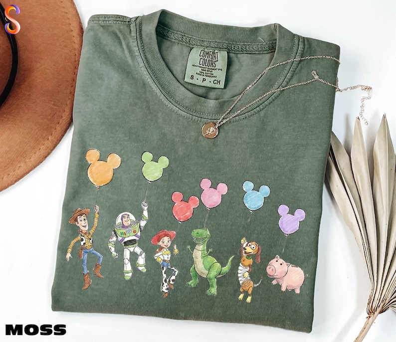 a green t-shirt with a design of various cartoon characters, including a character holding a balloon, a character holding a balloon, and several other characters, all depicted in a playful and colorful manner.