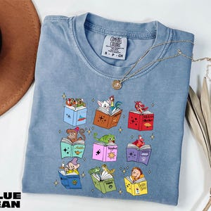 A blue denim t-shirt with a colorful graphic design of various cartoon characters and books, along with a necklace and a hat.
