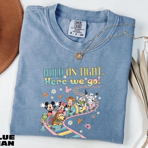 A blue denim t-shirt with a cartoon character design and text printed on it, along with a necklace and some decorative leaves.