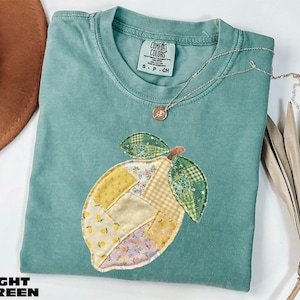 Puede incluir: Camiseta verde claro con un diseño de limón en patchwork. El limón está hecho de varias telas estampadas en amarillo, verde y rosa. Un collar plateado con un pequeño colgante descansa sobre la camisa. La camiseta tiene una etiqueta Comfort Colors.