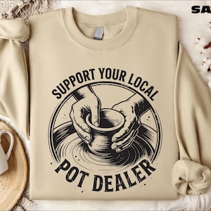 May include: Sand-colored sweatshirt with the text "SUPPORT YOUR LOCAL POT DEALER" in a black circular design. The design features hands shaping a pot on a pottery wheel. A white mug with a heart design is visible.