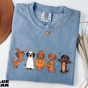 A blue t-shirt with a Halloween-themed graphic of five dogs dressed in costumes, including a witch, a pumpkin, a bat, a dog in a skeleton costume, and a dog in a skeleton costume holding a jack-o-lantern.