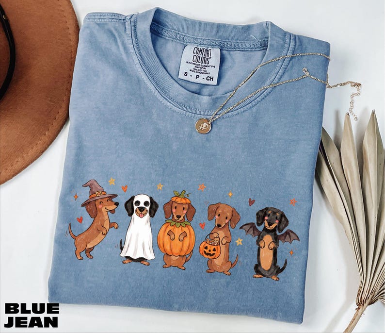 A blue t-shirt with a Halloween-themed graphic of five dogs dressed in costumes, including a witch, a pumpkin, a bat, a dog in a skeleton costume, and a dog in a skeleton costume holding a jack-o-lantern.