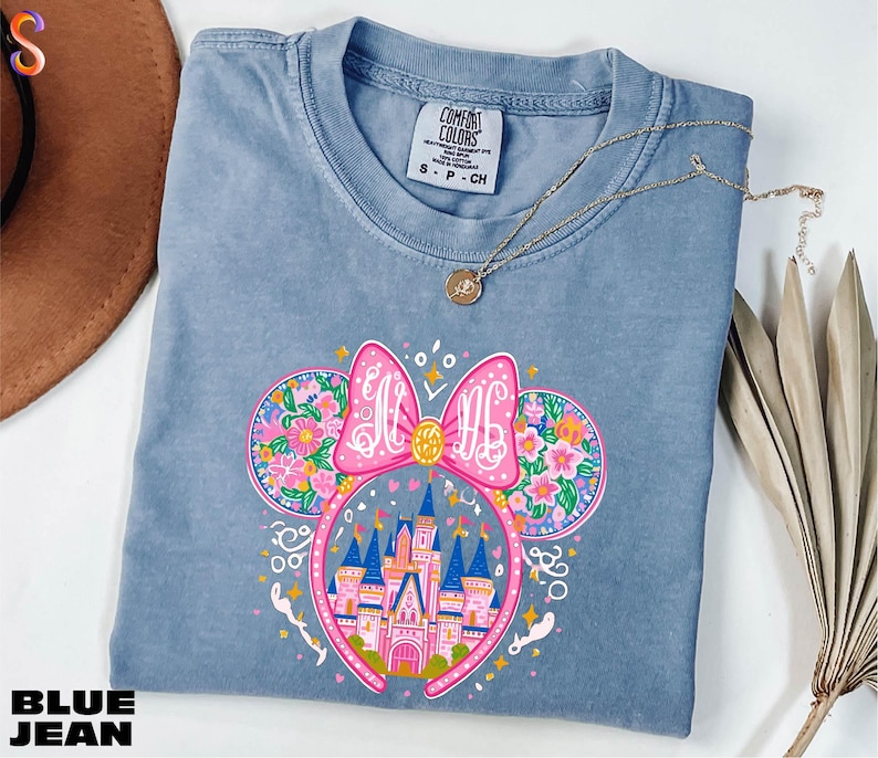 A blue t-shirt with a colorful graphic design featuring a castle and flowers, along with a necklace and a hat.