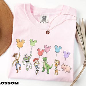 A pink t-shirt with a cartoon character design featuring a group of characters, including a boy, a girl, a dinosaur, a pig, and a bird, holding colorful balloons.