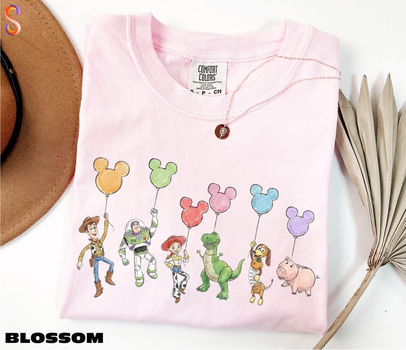 A pink t-shirt with a cartoon character design featuring a group of characters, including a boy, a girl, a dinosaur, a pig, and a bird, holding colorful balloons.