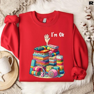 May include: Red sweatshirt with a white hand and the text "I'm Ok" printed on the front. The hand is holding a stack of colorful fabric scraps.