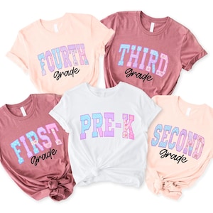 May include: Five short-sleeved t-shirts in shades of light pink, mauve, and white. Each shirt features a school grade level printed in a blue and pink patterned block font, with the word grade in black script below: Fourth, Third, First, Pre-K, Second.