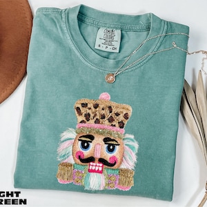 May include: A light green short-sleeve t-shirt featuring an embroidered nutcracker graphic with a leopard print hat, pink cheeks, and gold accents. The shirt has a Comfort Colors label at the neck and is paired with a delicate gold-tone necklace.