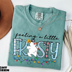 A light green t-shirt with a snowman graphic and the text "feeling a little FROSTY" printed on it, along with a necklace and a brown hat.