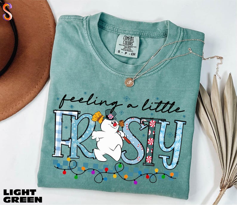 A light green t-shirt with a snowman graphic and the text "feeling a little FROSTY" printed on it, along with a necklace and a brown hat.
