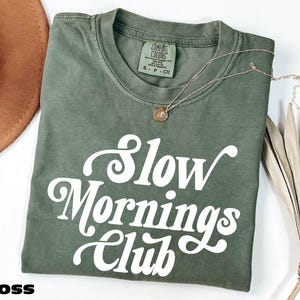 a green t-shirt with the text "Slow Mornings Club" printed on it, along with a necklace and a hat.