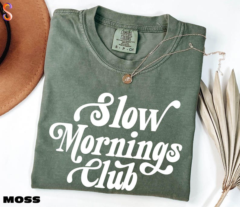 a green t-shirt with the text "Slow Mornings Club" printed on it, along with a necklace and a hat.