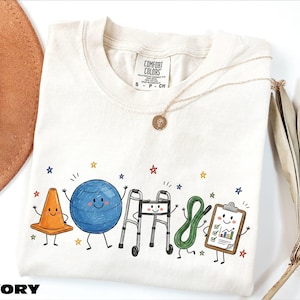 May include: An ivory t-shirt featuring a graphic print of five anthropomorphic physical therapy items: an orange traffic cone, a blue exercise ball, a silver walker, a green resistance band, and a clipboard with a chart, surrounded by small stars.