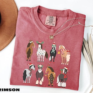 A red t-shirt with a graphic of nine horses wearing Christmas-themed outfits, including Santa hats and scarves, is displayed on a white surface alongside a brown hat and some greenery.