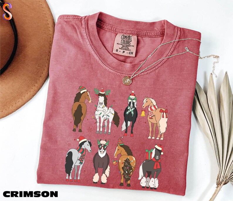 A red t-shirt with a graphic of nine horses wearing Christmas-themed outfits, including Santa hats and scarves, is displayed on a white surface alongside a brown hat and some greenery.