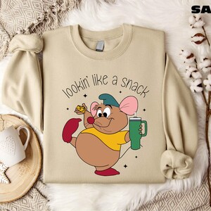 K&ouml;nnte beinhalten: Sandfarbenes Sweatshirt mit einer Cartoon-Maus, die K&auml;se und einen gr&uuml;nen Becher h&auml;lt. Die Maus tr&auml;gt ein gelbes Hemd, rote Schuhe und einen blauen Hut. Der Text "lookin' like a snack" steht &uuml;ber der Maus.