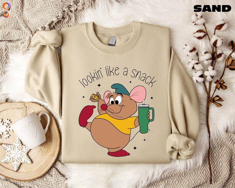 K&ouml;nnte beinhalten: Sandfarbenes Sweatshirt mit einer Cartoon-Maus, die K&auml;se und einen gr&uuml;nen Becher h&auml;lt. Die Maus tr&auml;gt ein gelbes Hemd, rote Schuhe und einen blauen Hut. Der Text "lookin' like a snack" steht &uuml;ber der Maus.