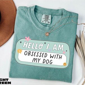May include: Light green t-shirt with the text "HELLO I AM OBSESSED WITH MY DOG" in a white and black design. The shirt has a crew neck and short sleeves. A silver necklace with a pendant is on the shirt.