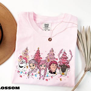 A pink t-shirt with a Christmas-themed design featuring characters from the Disney movie Frozen, including Elsa, Anna, Olaf, and a reindeer. The t-shirt is displayed on a white background, along with a hat, a necklace, and a palm frond.