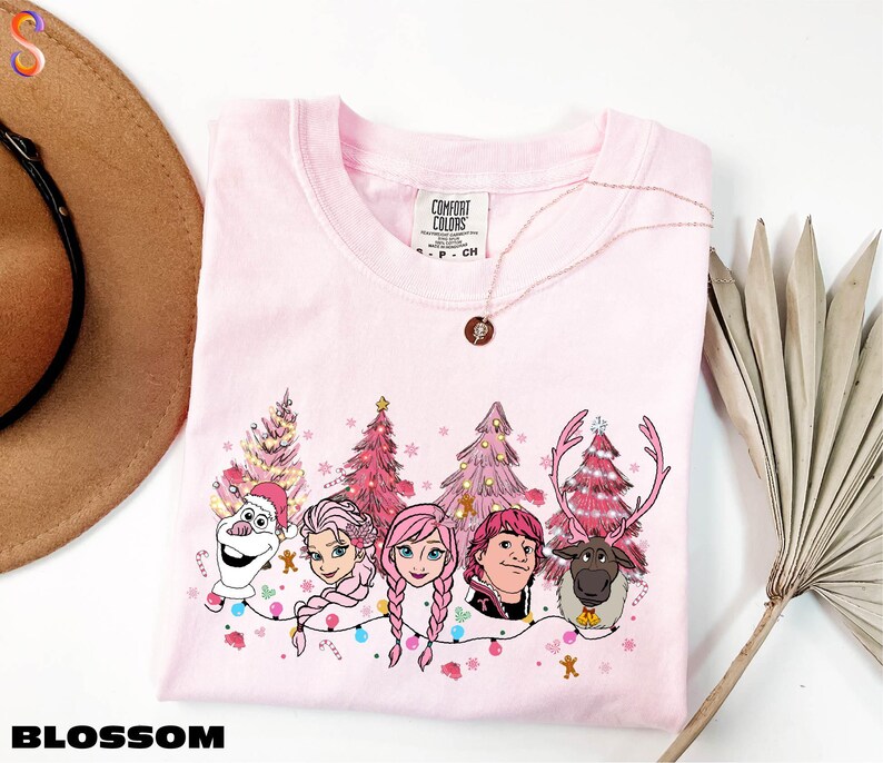 A pink t-shirt with a Christmas-themed design featuring characters from the Disney movie Frozen, including Elsa, Anna, Olaf, and a reindeer. The t-shirt is displayed on a white background, along with a hat, a necklace, and a palm frond.