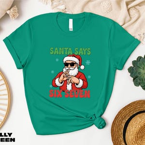 Santa Claus wearing sunglasses and holding up his hand with fingers in front of his face, wearing a green t-shirt with the text "SANTA SAYS SIX SEVEN" printed on it.