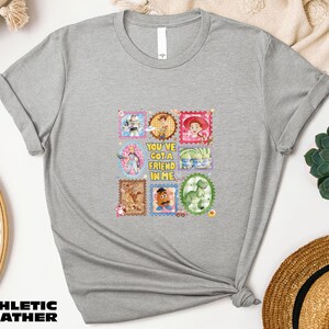 a gray t-shirt with a colorful graphic design, including various cartoon characters and text. The t-shirt is placed on a white surface, surrounded by other items such as a hat, a potted plant, and a woven basket.