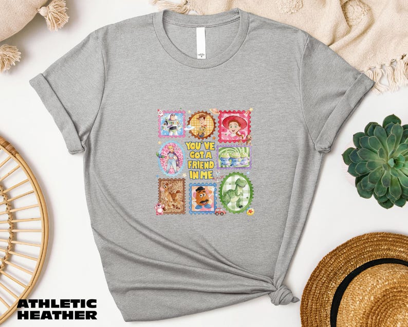 a gray t-shirt with a colorful graphic design, including various cartoon characters and text. The t-shirt is placed on a white surface, surrounded by other items such as a hat, a potted plant, and a woven basket.