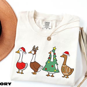 A white t-shirt with a Christmas-themed design featuring four cartoon ducks in festive attire, including Santa hats and Christmas trees.