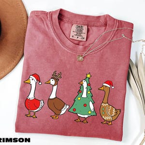 A pink t-shirt with a Christmas-themed graphic of four cartoon ducks in festive attire, including Santa Claus, reindeer, and a Christmas tree.