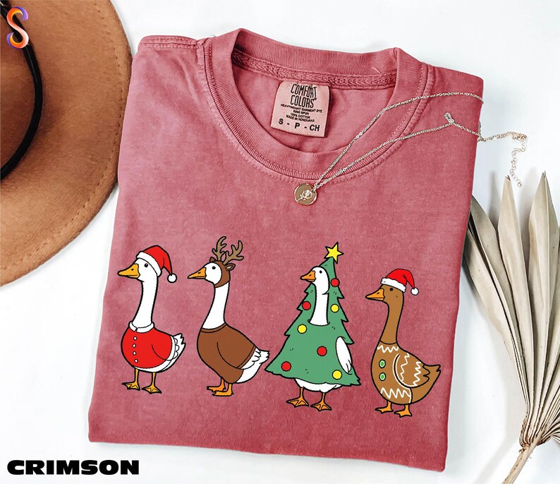A pink t-shirt with a Christmas-themed graphic of four cartoon ducks in festive attire, including Santa Claus, reindeer, and a Christmas tree.