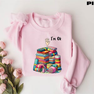 May include: Pink sweatshirt with a graphic of a pile of colorful fabric scraps and the text "I'm Ok".