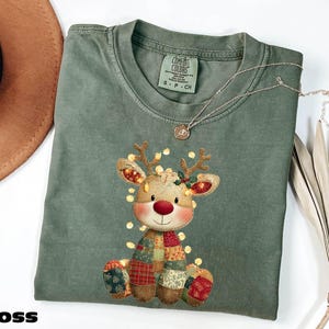 Peut inclure: Un t-shirt vert mousse Comfort Colors avec un motif de renne festif. Le renne a un corps en patchwork, un nez rouge et est orné de guirlandes lumineuses. Le mot "MOSS" est imprimé en gras, en noir en bas.