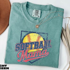 A light green t-shirt with a graphic of a softball and the text "SOFTBALL Mama" printed on it, along with a necklace and a brown hat.