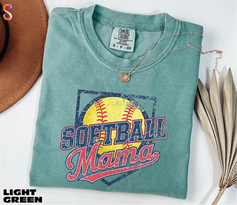 A light green t-shirt with a graphic of a softball and the text "SOFTBALL Mama" printed on it, along with a necklace and a brown hat.