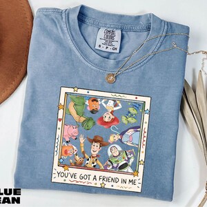 A blue denim t-shirt with a cartoon character design and text, along with a necklace and a hat, all placed on a white surface.