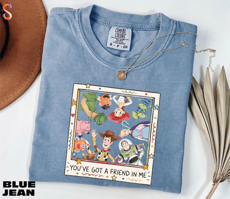 A blue denim t-shirt with a cartoon character design and text, along with a necklace and a hat, all placed on a white surface.