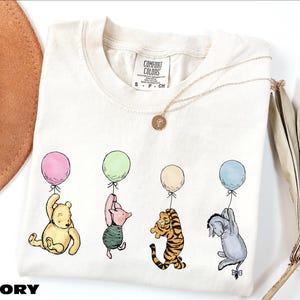 A white t-shirt with a printed design of Winnie the Pooh and other characters holding balloons, along with a necklace and a hat.