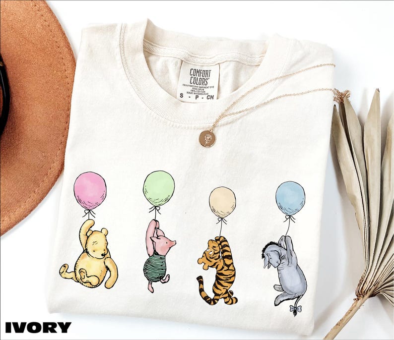 A white t-shirt with a printed design of Winnie the Pooh and other characters holding balloons, along with a necklace and a hat.