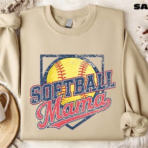 A beige softball-themed sweatshirt with the text "SOFTBALL MAMA" printed on it, along with a baseball graphic and the word "MAMA" written in cursive. The sweatshirt is laid out on a surface with a mug and some cotton balls.