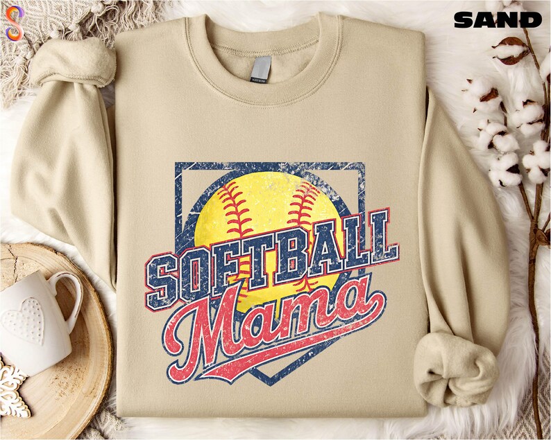 A beige softball-themed sweatshirt with the text "SOFTBALL MAMA" printed on it, along with a baseball graphic and the word "MAMA" written in cursive. The sweatshirt is laid out on a surface with a mug and some cotton balls.