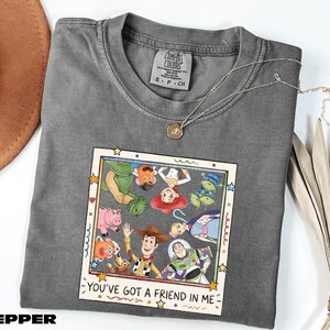 A gray t-shirt with a graphic design featuring characters from the Toy Story movie, including Woody, Buzz Lightyear, and other beloved characters. The text on the shirt reads "You've Got a Friend in Me".