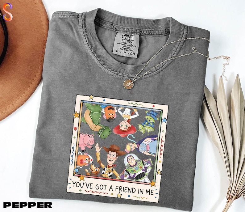 A gray t-shirt with a graphic design featuring characters from the Toy Story movie, including Woody, Buzz Lightyear, and other beloved characters. The text on the shirt reads "You've Got a Friend in Me".