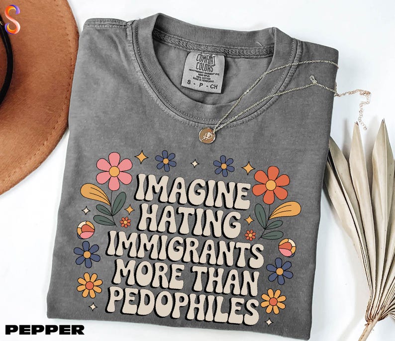 A gray t-shirt with a colorful graphic design and text printed on it, along with a necklace and some dried flowers.