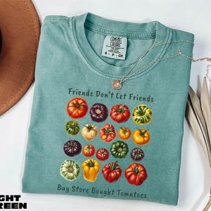 A light blue t-shirt with a graphic design of various colorful tomatoes, accompanied by the text "Friends Don't Let Friends" and "Buy Store Bought Tomatoes".