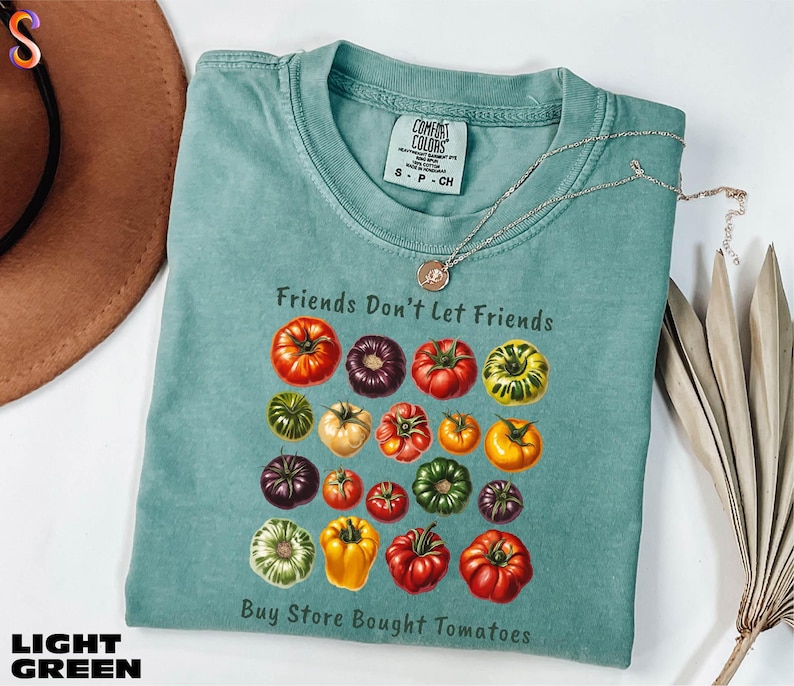 A light blue t-shirt with a graphic design of various colorful tomatoes, accompanied by the text "Friends Don't Let Friends" and "Buy Store Bought Tomatoes".