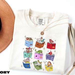 A white t-shirt with a colorful graphic design of various cartoon characters, including a dragon, a unicorn, and other whimsical figures, along with some books and a hat.