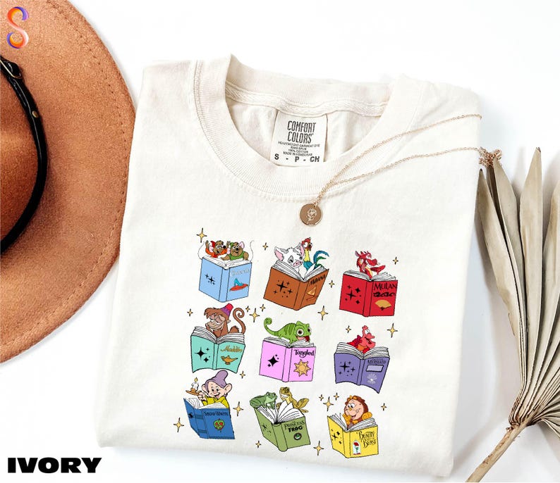 A white t-shirt with a colorful graphic design of various cartoon characters, including a dragon, a unicorn, and other whimsical figures, along with some books and a hat.