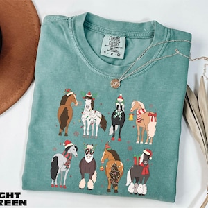 A turquoise t-shirt with a graphic of nine horses wearing Christmas-themed outfits, including Santa hats and scarves, is displayed on a white surface alongside a brown hat and some dried leaves.