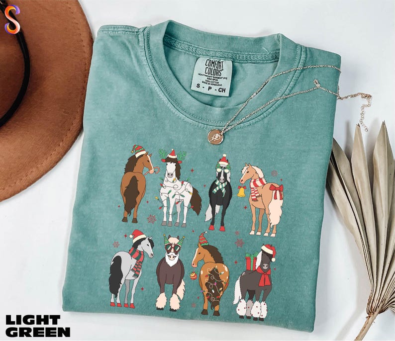 A turquoise t-shirt with a graphic of nine horses wearing Christmas-themed outfits, including Santa hats and scarves, is displayed on a white surface alongside a brown hat and some dried leaves.